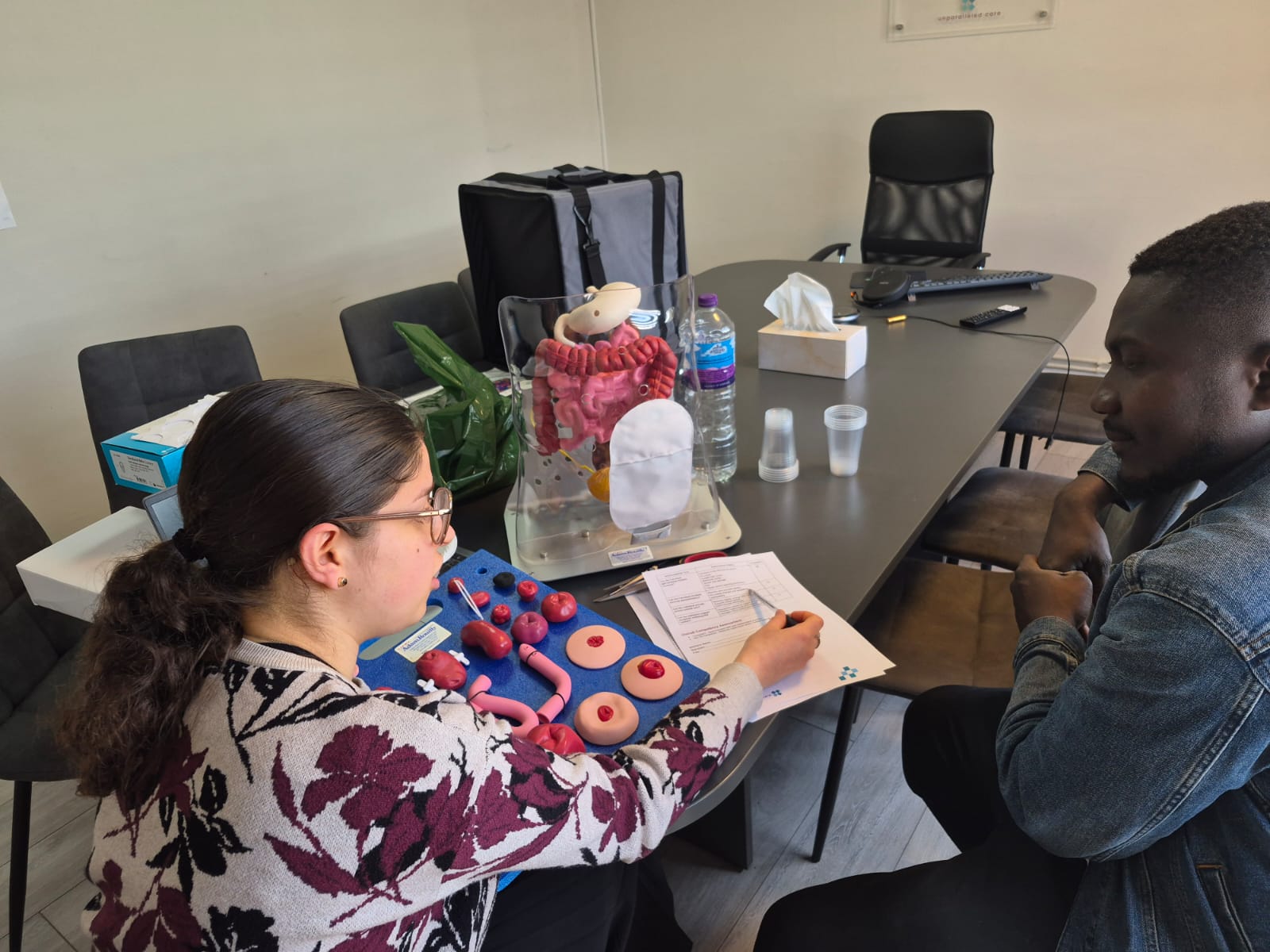 A woman showing care training equipment with an anatomical model to a seated man across a table.